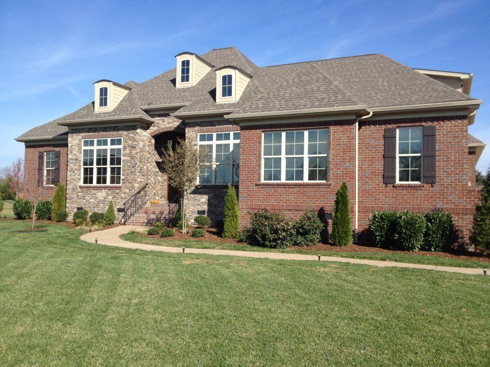 Suburban brick house with manicured lawn, stone accents, and dormer windows under a blue sky