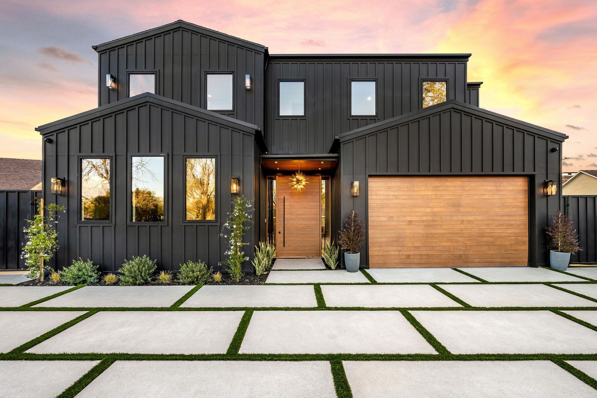 Modern two-story dark gray house with wood garage, lit windows, and a geometric concrete driveway at sunset