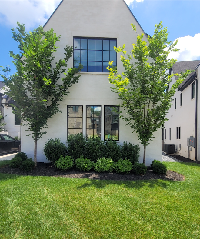 White two-story house with large windows, trimmed shrubs, and a green lawn under a blue sky.