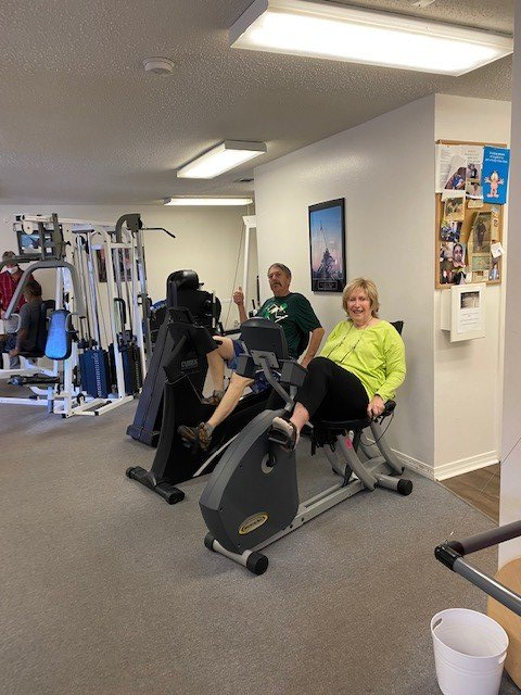 People using gym equipment in a bright fitness room, with a white bucket in the foreground.