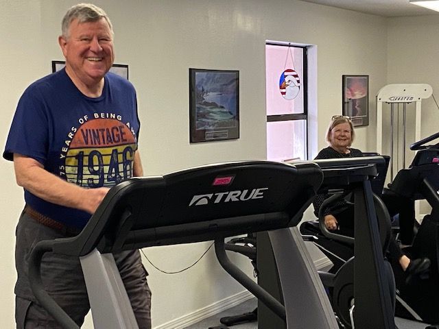 Two people smiling on treadmills in a gym, one in a blue shirt in front and one seated behind.