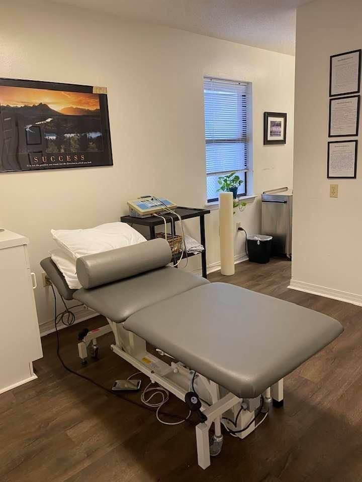 Massage table in a small treatment room with framed art, window, and supply cart.