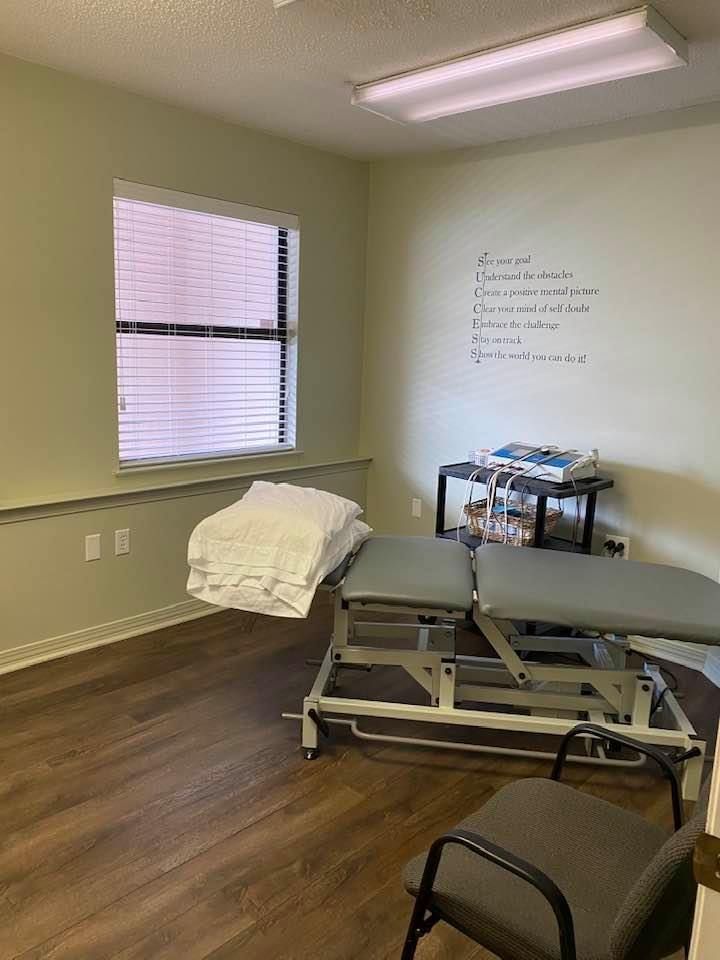 Exam room with treatment table, stool, chair, window, and whiteboard on beige walls.