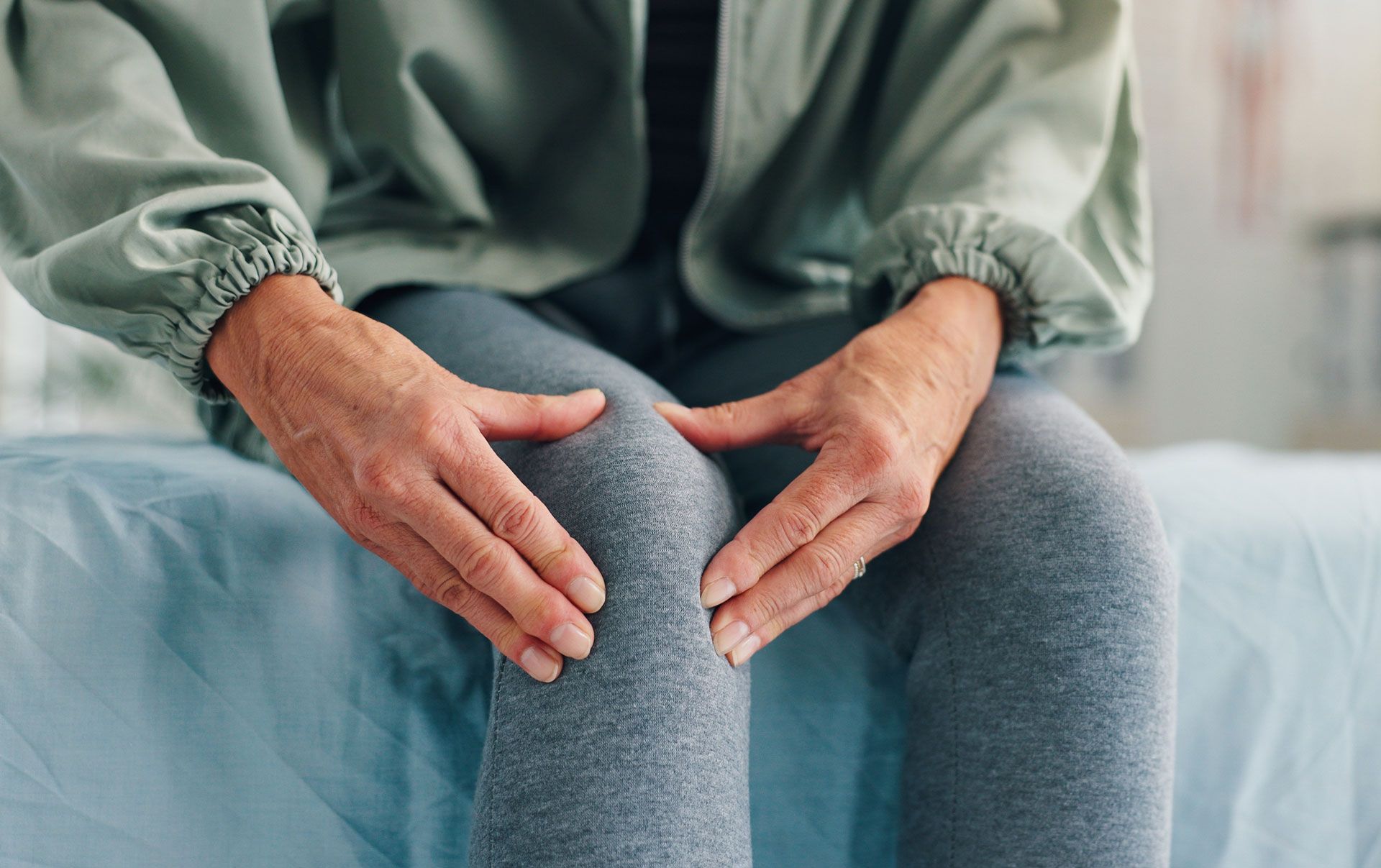 Hands resting on knees while seated on a bed, suggesting knee pain or discomfort.