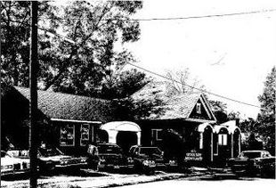Black-and-white photo of a house with cars parked outside under trees and power lines.