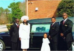 Four people stand beside a black hearse outside a brick building, with a child in a white dress in front.