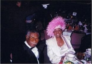 Two people seated at a formal event, one wearing a white suit and pink feathered hat.