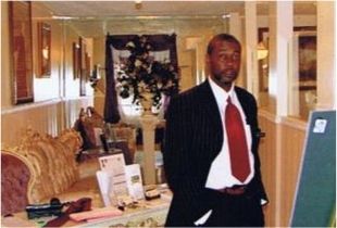 Man in suit and red tie standing in a hotel lobby beside a display board