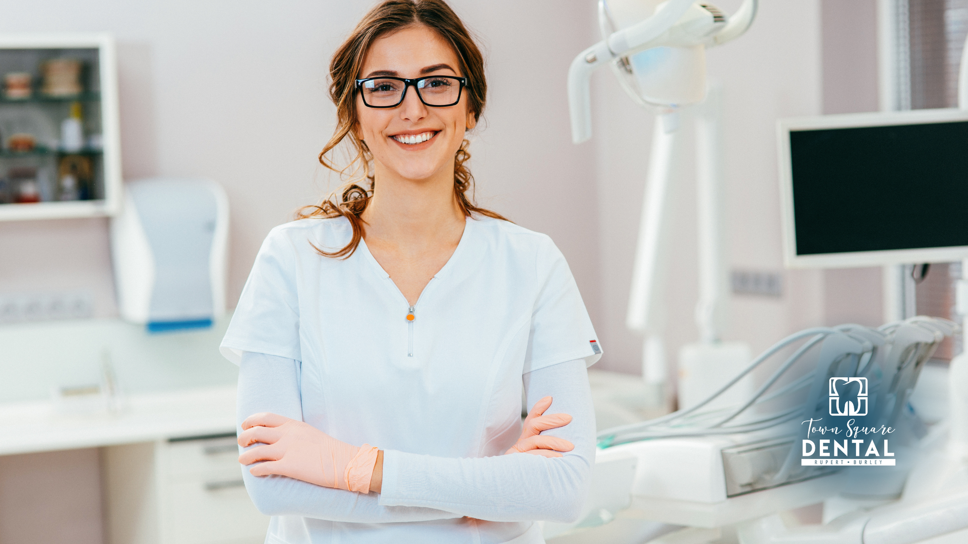 A female dentist is standing in a dental office with her arms crossed and smiling.