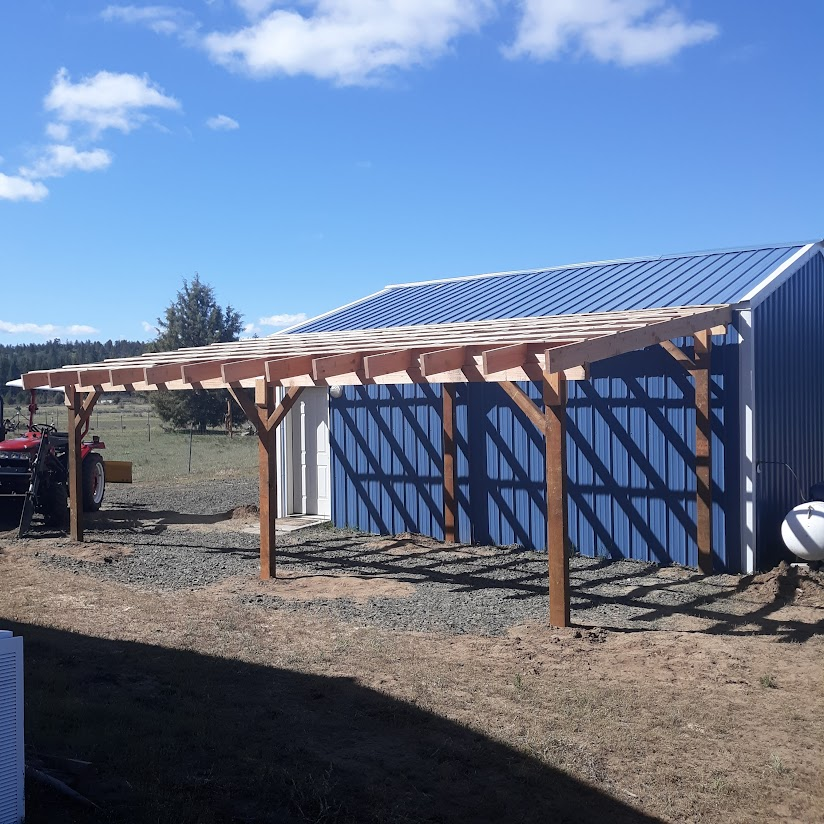 Wooden carport built next to a blue metal-sided shed under a blue sky.