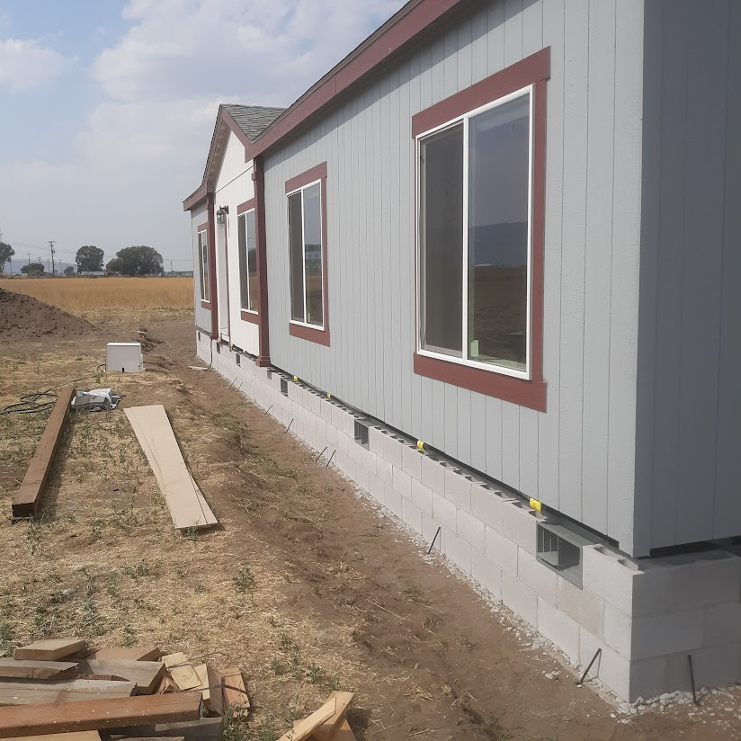 Side view of a light gray mobile home with dark red trim and white windows, on a concrete block foundation.