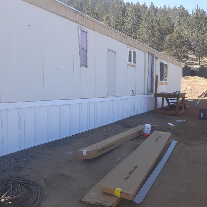 Mobile home with white siding and skirting; construction materials are in foreground.