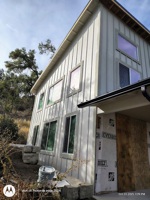 Modern house with light gray vertical siding and multiple windows on a hillside, under construction.