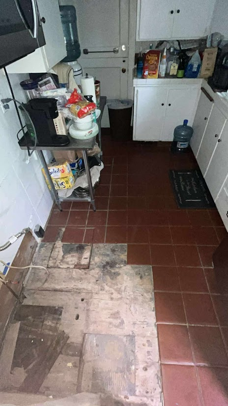Kitchen with damaged floor tiles and exposed subfloor. White cabinets, water cooler, and miscellaneous items visible.