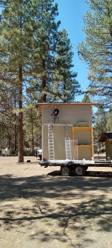 Person standing on the roof of a small building on a trailer in a wooded area.