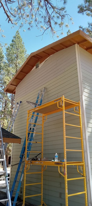 A building with a wooden roof and light yellow siding, with scaffolding and a ladder alongside.