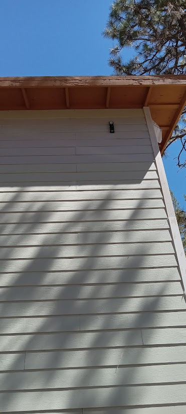 Side of a light-colored building with horizontal siding. Brown roof and a blue sky above. Shadows from a tree.