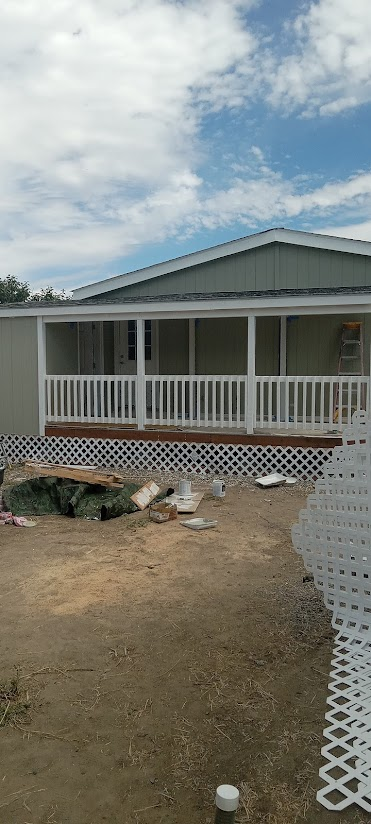 A house with a porch and white railings sits on dirt ground with scattered debris under a cloudy sky.