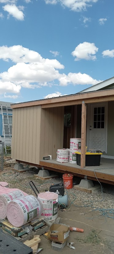 Exterior of a building under construction, with a porch. Materials are on the ground and porch. Blue sky with clouds.