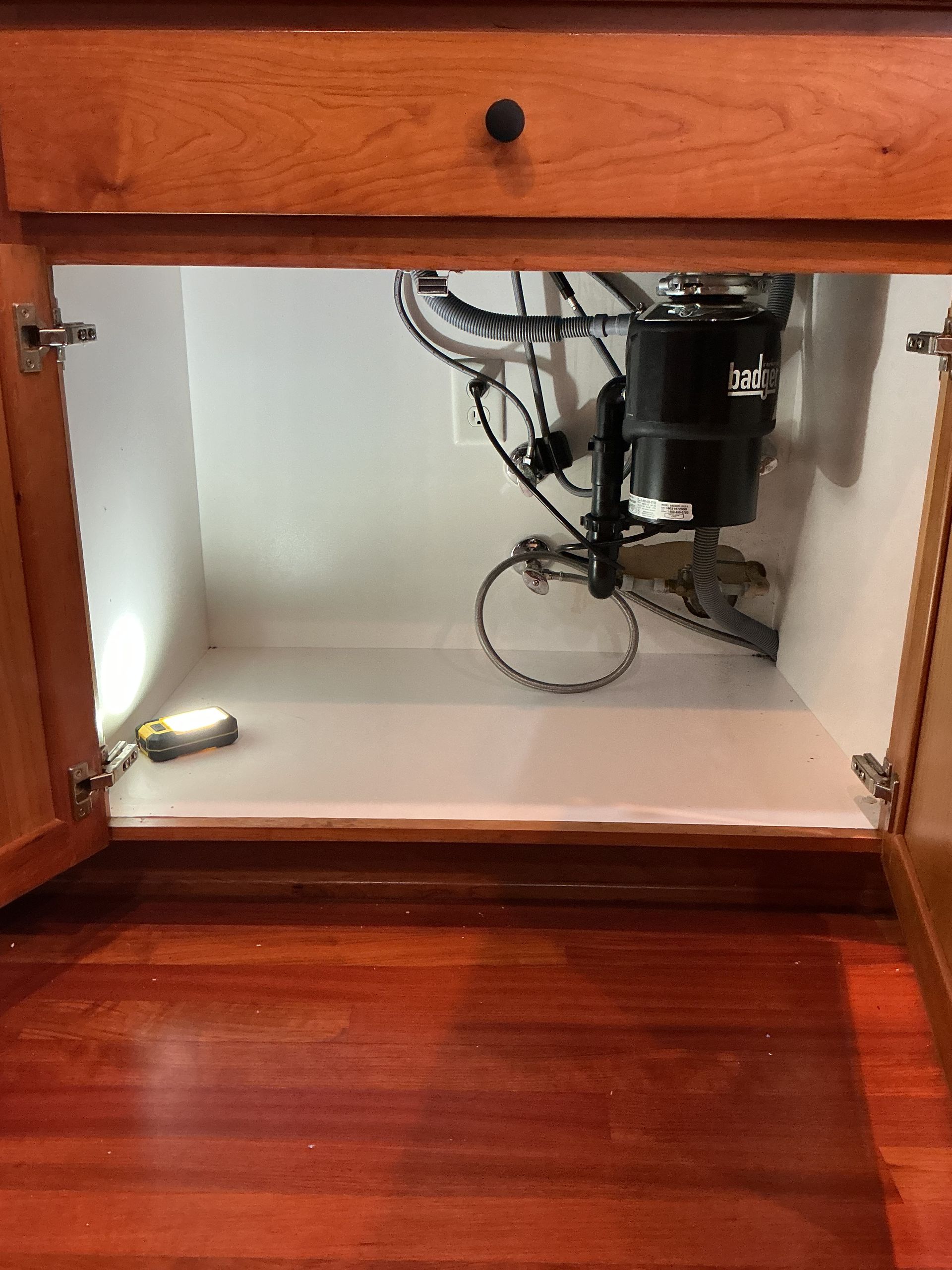 Open cabinet under a sink with exposed plumbing and a flashlight inside, white interior and wooden door.