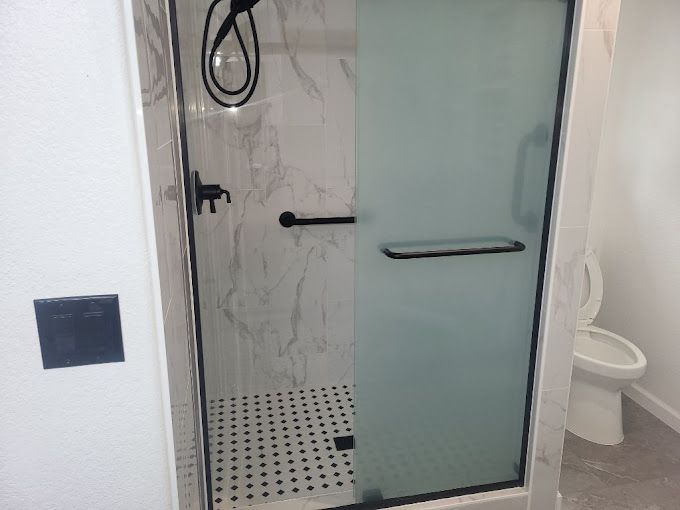 Small bathroom shower with frosted glass door, white tile, black grab bars, and a toilet beside it.