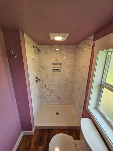 Small pink bathroom with marble shower, window, toilet, and wood-look floor.