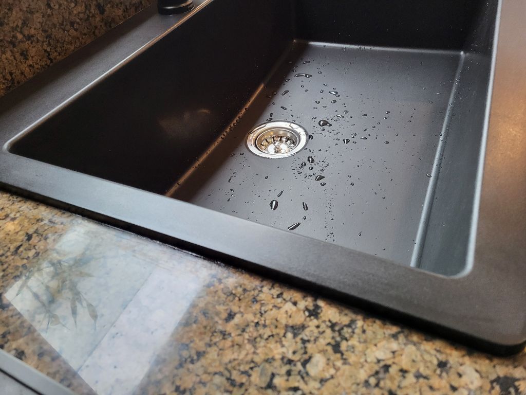 Black kitchen sink with silver drain and water droplets on a granite countertop
