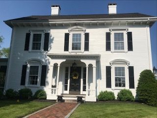 A large white house with black shutters and a brick walkway leading to the front door.