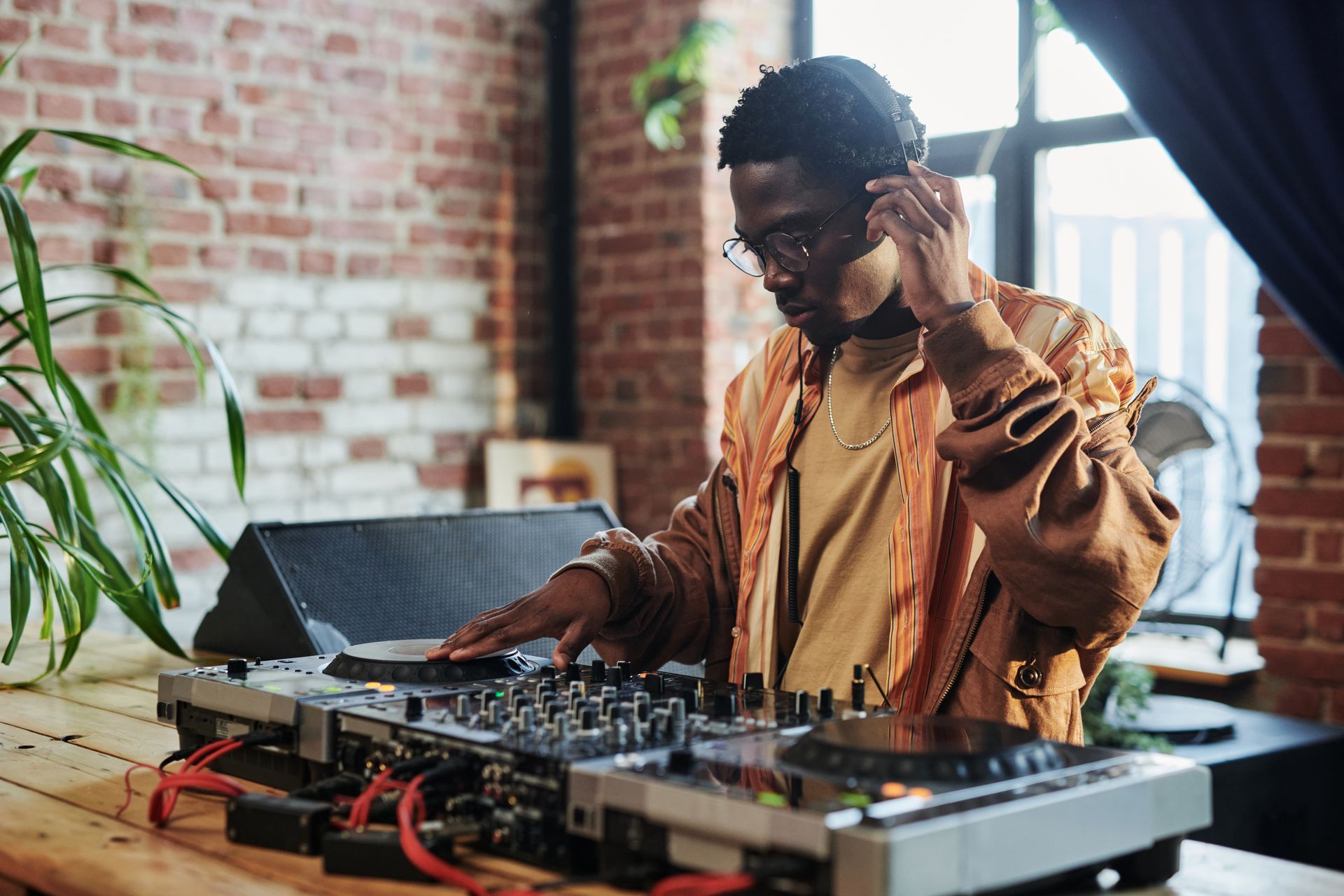 DJ mixing music at a table with turntables and headphones in front of a brick wall.