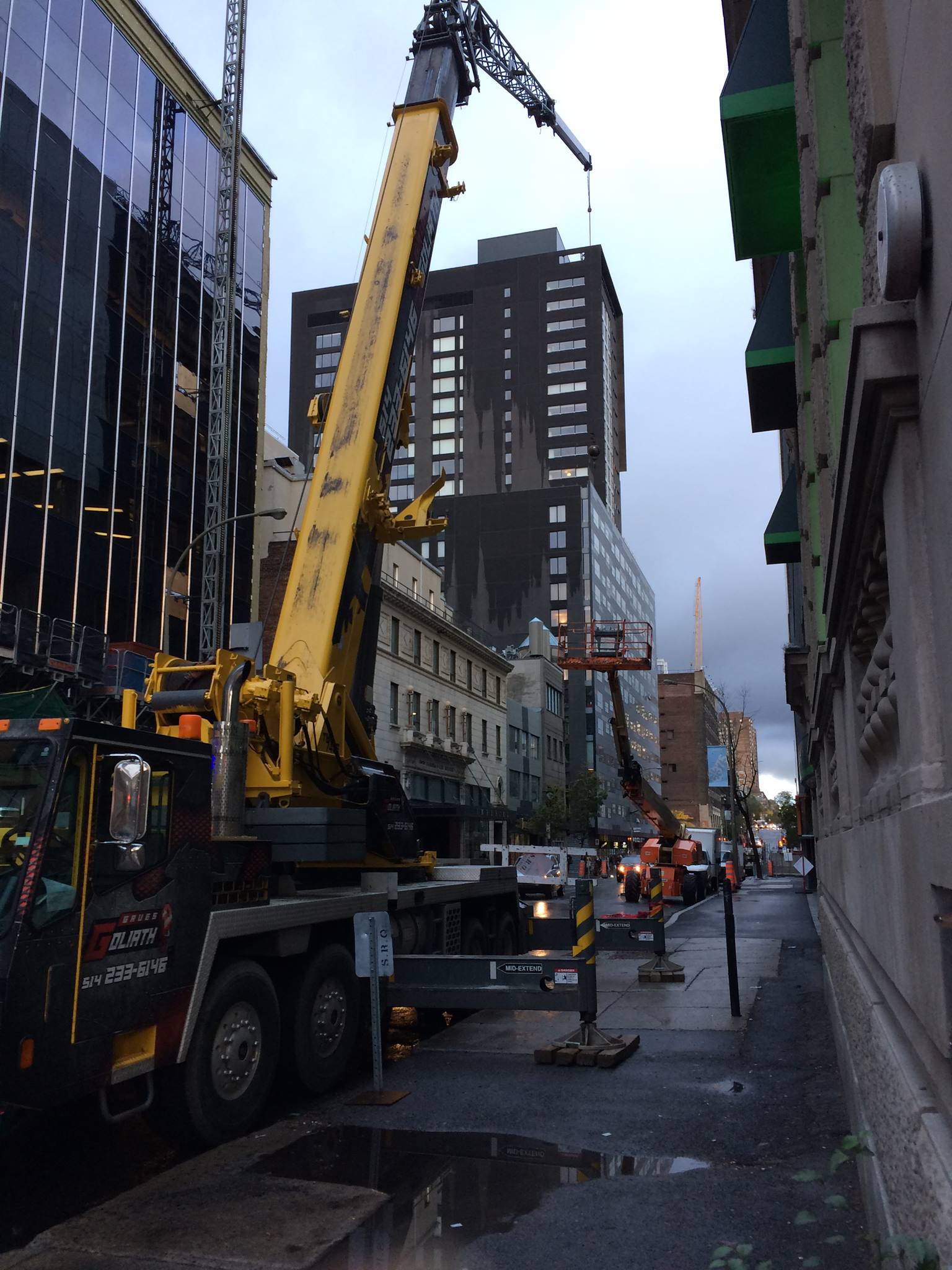 Une grande grue jaune soulevant des matériaux dans une rue de la ville, avec de grands immeubles en arrière-plan.