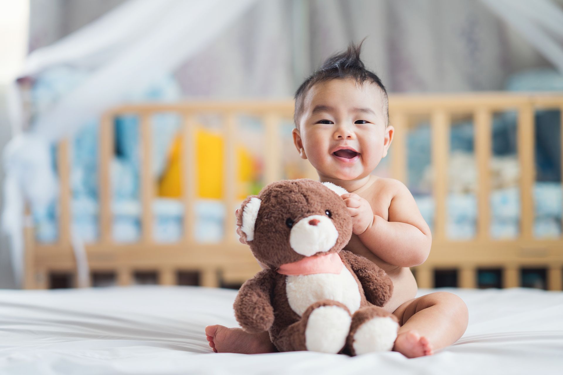 Smiling baby with mohawk sits on a bed, cuddling a brown teddy bear in front of a crib.