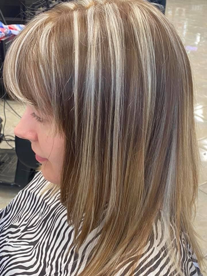 Woman with shoulder-length hair, highlights of blonde and brown. She wears a patterned smock, in a salon setting.