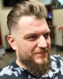 Man with blonde pompadour, faded sides, and beard, smiling indoors.