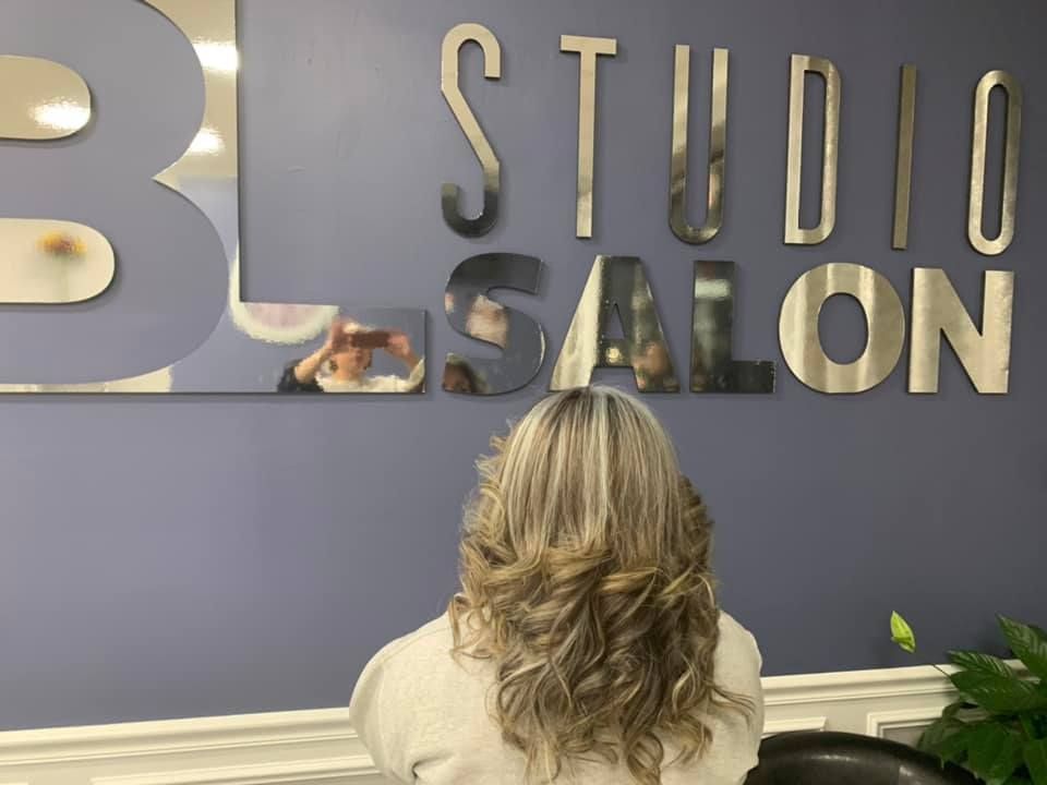Woman with wavy, layered blonde hair in a salon, in front of a wall sign that says