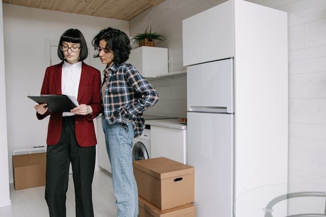 woman in red blazer holding clipboard showing it to another woman in a plaid shirt in a small white apartment