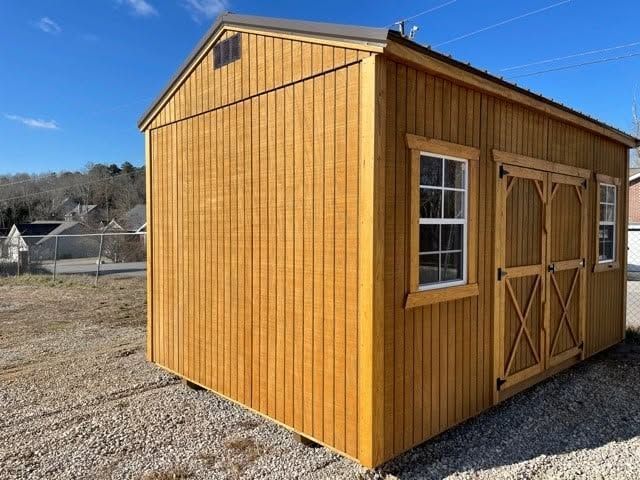 Small yellow wooden shed with a gambrel roof and double doors on a gravel lot