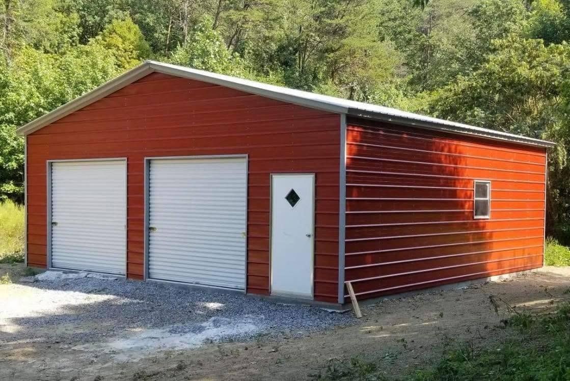 Red detached garage with two white doors and a side entrance, set on a gravel drive near trees.