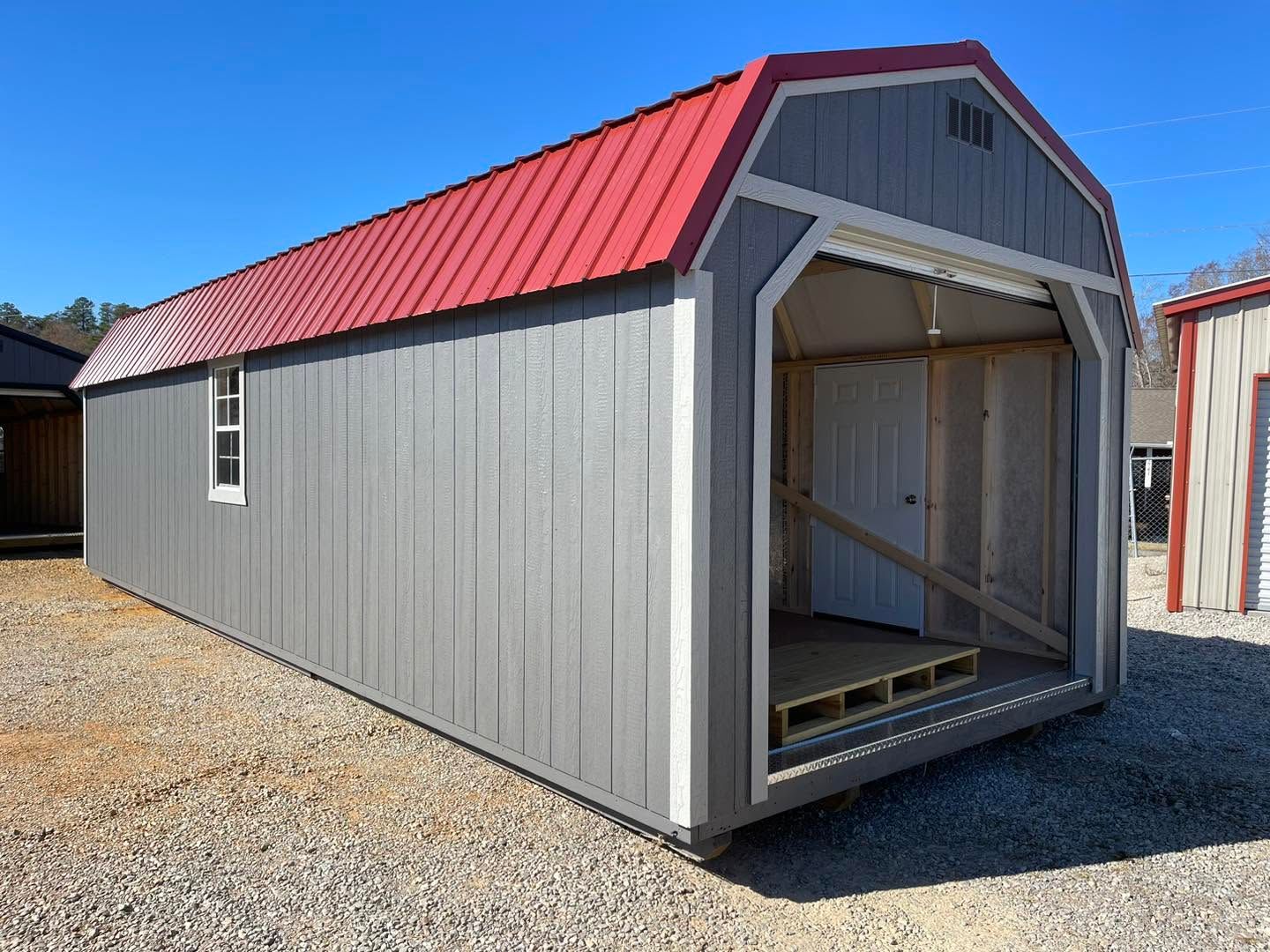 Small gray shed with red metal roof and open front on a gravel lot
