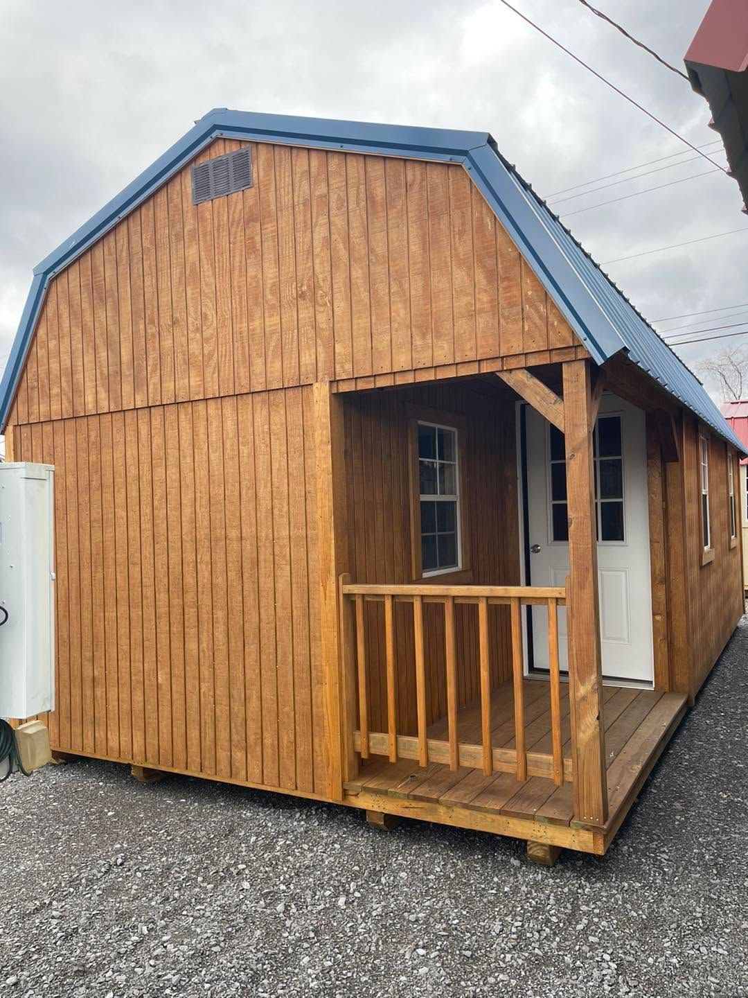 Small wooden cabin with blue roof and tiny porch on gravel, viewed from the front corner