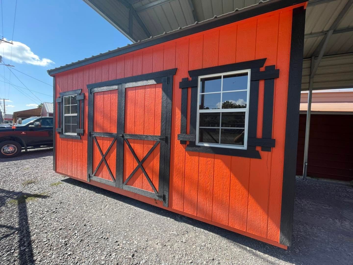 Orange-red shed with black-trimmed double doors and a window on a gravel lot under a metal roof.