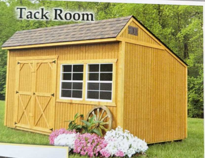 Yellow wooden tack room shed with double doors, windows, wagon wheel, and flowers on grass.