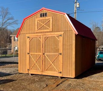Large wooden barn with red roof and double doors on a gravel lot under a clear blue sky