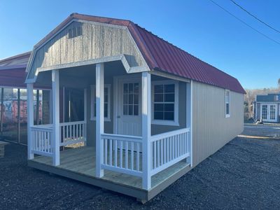 Small beige cottage with red roof and white porch railings on a gravel lot.