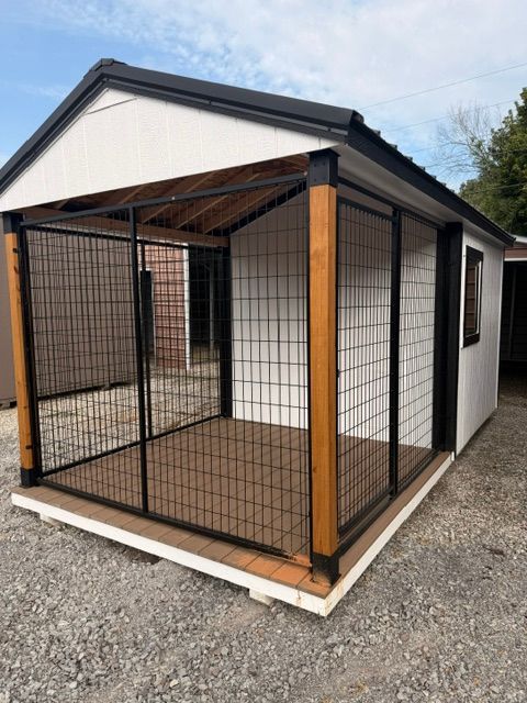 Small fenced kennel with black mesh panels and a slanted roof beside a white outbuilding.