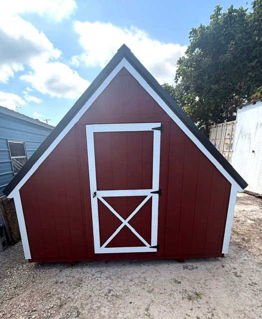 Red wooden shed with white trim and X-braced door, sitting on gravel outdoors under a blue sky.