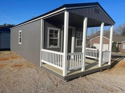 Small gray shed-style porch with white railings and a covered front entry on a gravel lot.