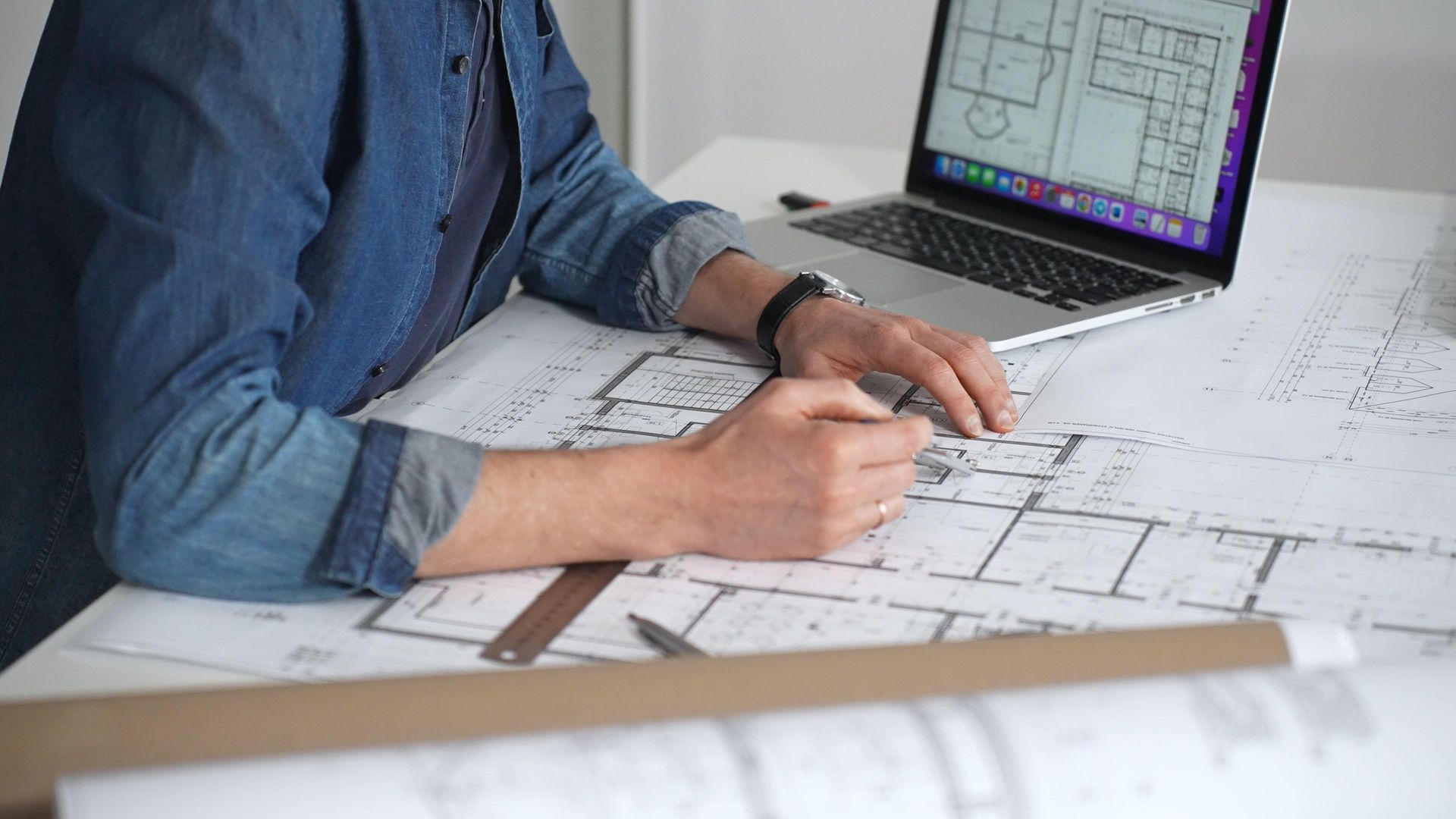 Person in a denim shirt sketches architectural plans beside a laptop displaying floor plans.