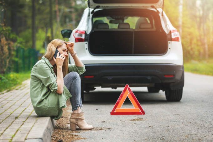 Woman sitting by broken-down SUV on roadside, holding phone and looking worried, with warning triangle nearby