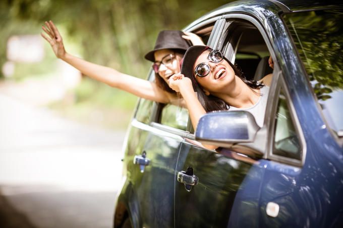 Two smiling people leaning out of a blue car window, waving on a sunny road