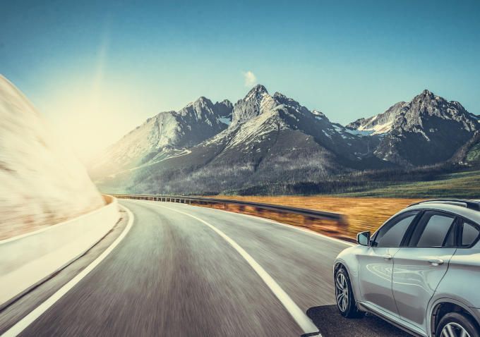Silver car driving on a curving mountain road with snowy peaks in the distance and bright sunlight on the left